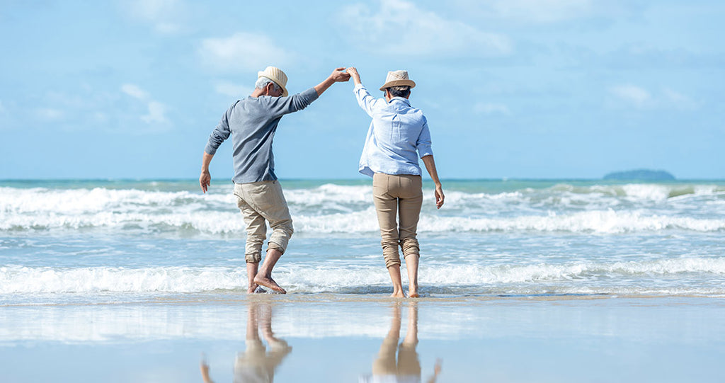 Older couple hold hands on beach and walk barefoot into the ocean on a clear day