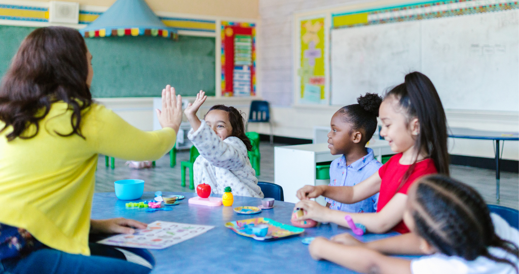 A classroom scene during back-to-school season. A teacher in a yellow sweater gives a high-five to an excited young student. 