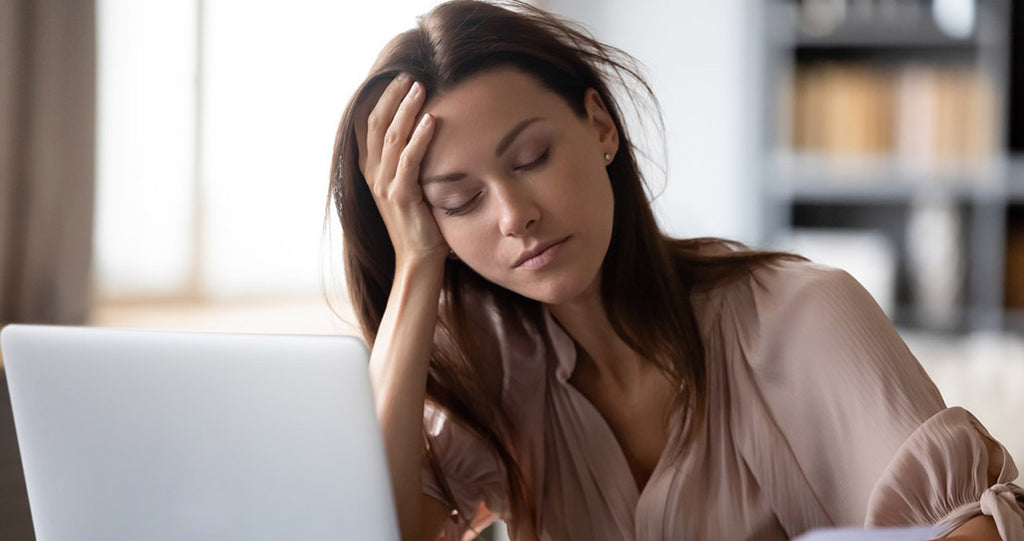 A woman with long dark hair sits at a desk, looking tired and stressed. She has her hand on her forehead and eyes closed, with a laptop in front of her.