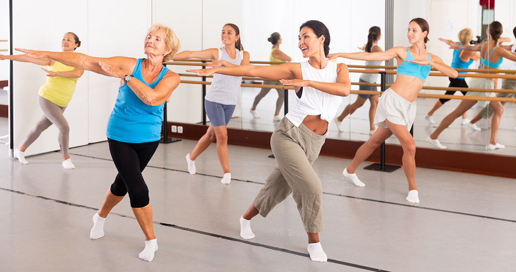 A group of women in a fitness class lunge with their arms stretched to their sides enjoying some sort of zumba or dance instruction that is getting their heart rate up and improving their overall health