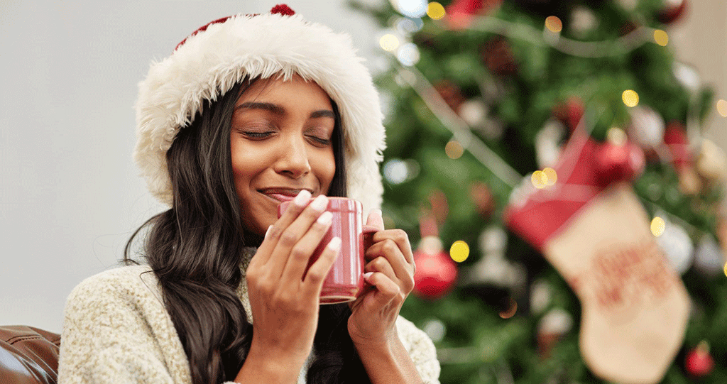 A woman wearing a cozy white winter hat and sweater sitting in front of a Christmas tree, holding a warm mug and smiling contentedly with her eyes closed.