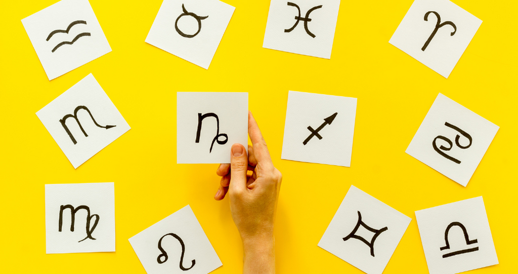 On a bright yellow background, multiple white squares are arranged, each containing a hand-drawn zodiac symbol. A person's hand is visible, holding one of the squares with the Capricorn symbol. The layout suggests a playful, astrological theme.