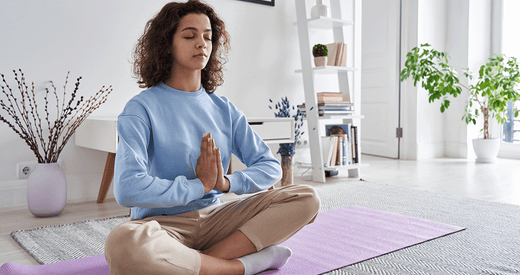 Woman in blue sweatshirt sitting cross-legged on yoga mat practicing meditation in bright, minimalist home space.