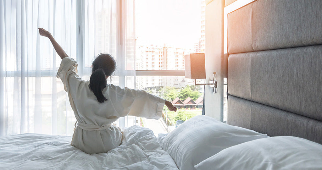 A person in a white bathrobe stretches their arms wide while sitting on  a bed, facing a sunlit window that overlooks a city skyline.