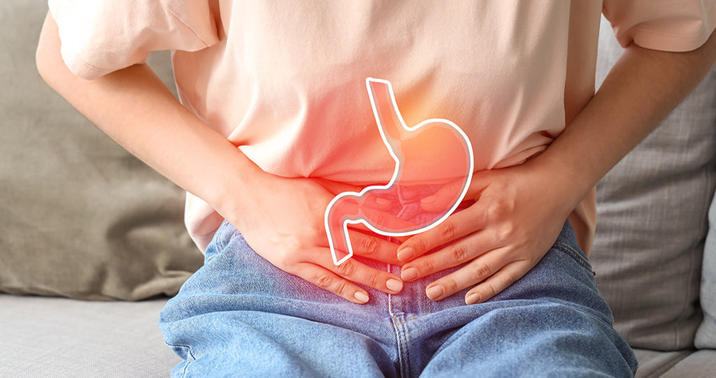 Person sitting on a bed, wearing a light pink shirt and blue jeans. They are holding their stomach with both hands. An outline of a stomach is superimposed on their abdomen, glowing red to indicate pain or discomfort. 