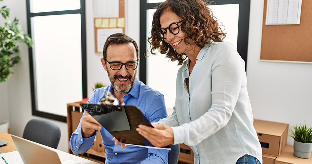 Two coworkers review information on a tablet in a bright office space. A seated man holds the device while a woman leans in, pointing at the screen.